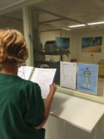 Thumbnail of A buddy nurse demonstrates reading instructions in front of the isolation unit glass window for healthcare personnel working inside the unit, Major Incident Hospital, University Medical Centre of Utrecht, the Netherlands, 2014.