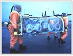 Thumbnail of Aeromedical isolation team members in field-protective suits equipped with battery-powered HEPA-filtered respirators transporting the stretcher isolator, a light-weight unit designed for initial patient retrieval. The team trains on several types of military aircraft, including the C-130 transport shown in the background.